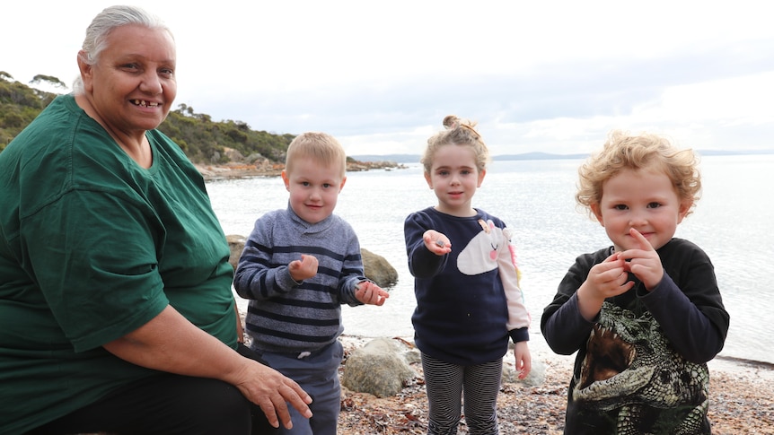 Indigenous woman sitting on rock with three young children holding shells at a rocky beach, headland in background