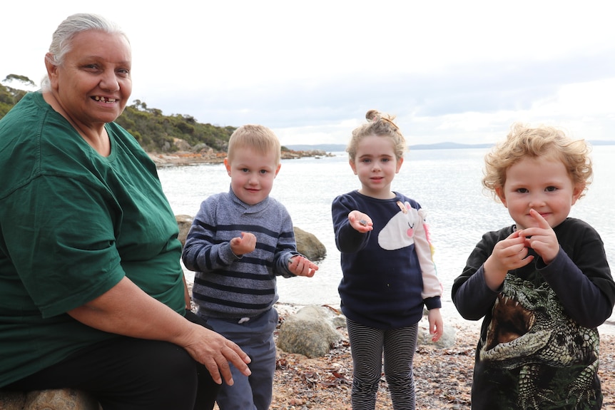 Indigenous woman sitting on rock with three young children holding shells at a rocky beach, headland in background