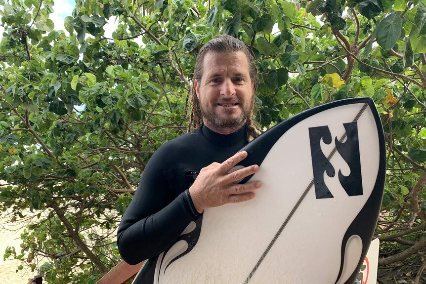 Surf legend Mark Occhilupo stares at camera holding a surfboard.