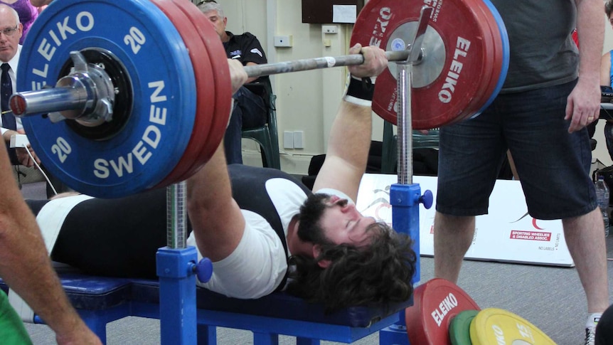 A man lying on a bench doing a bench press with weights while another man watches over and spectators watch in the background