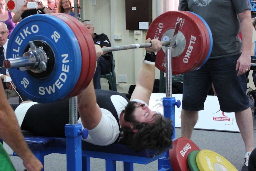 A man lying on a bench doing a bench press with weights while another man watches over and spectators watch in the background