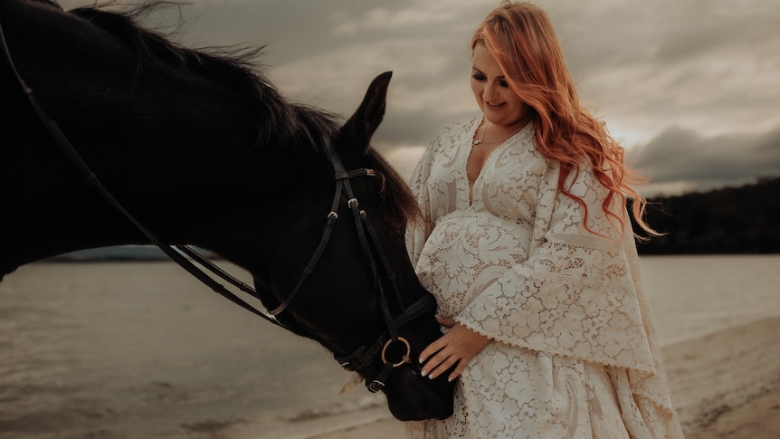 A pregnant woman in a white dress with a horse on a beach.