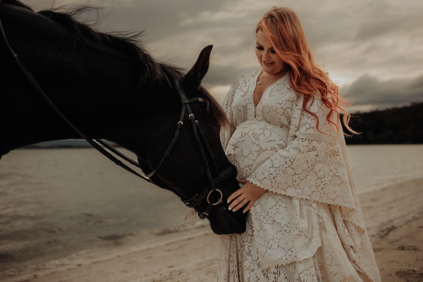 A pregnant woman in a white dress with a horse on a beach.