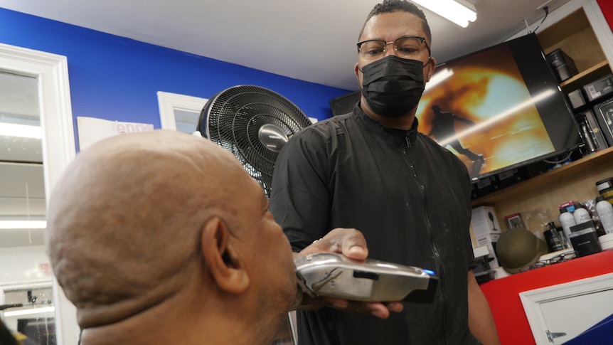 a man with a black face mask and a black shirt and glasses holds an electric razor to a sitting man's face