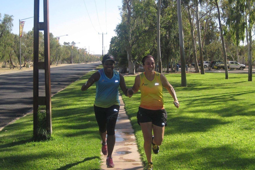 Two smiling female marathon runners hold hands and run down a suburban street.