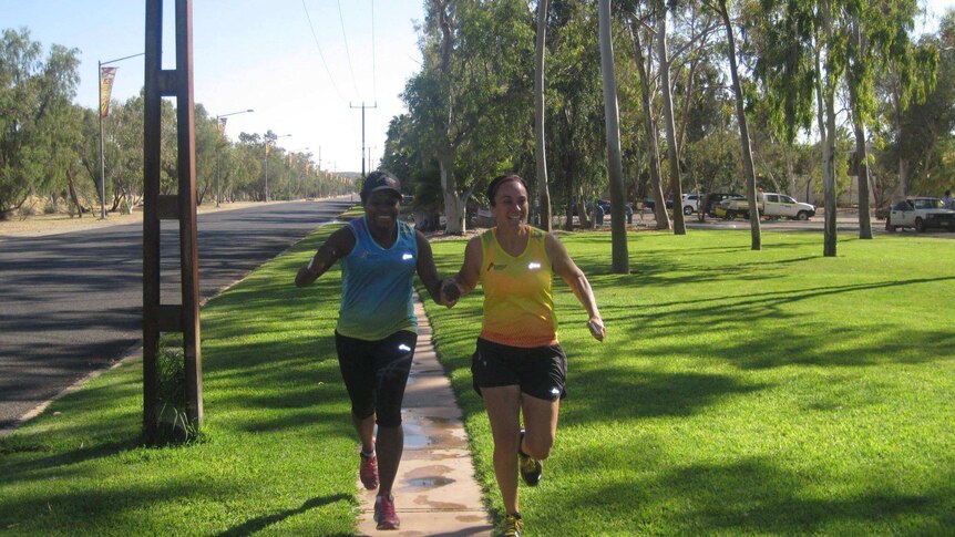 Two smiling female marathon runners hold hands and run down a suburban street.