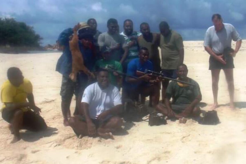 A group of men stand on an island sandy beach. One person holds a dead goat. 