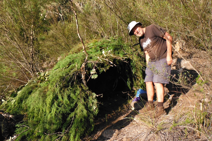 A middle-aged man poses next to a shelter built out of ferns.