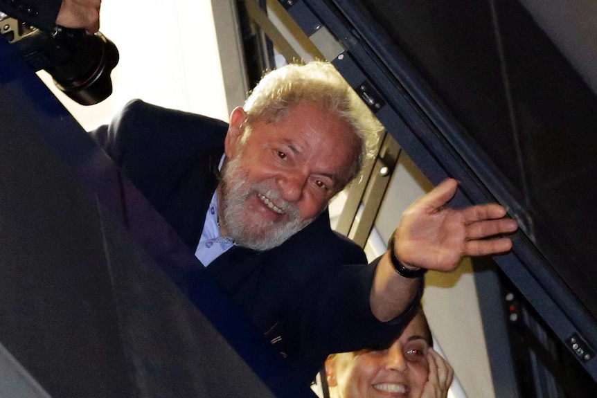 Man waves his left hand, smiling. Woman behind him also smiles.