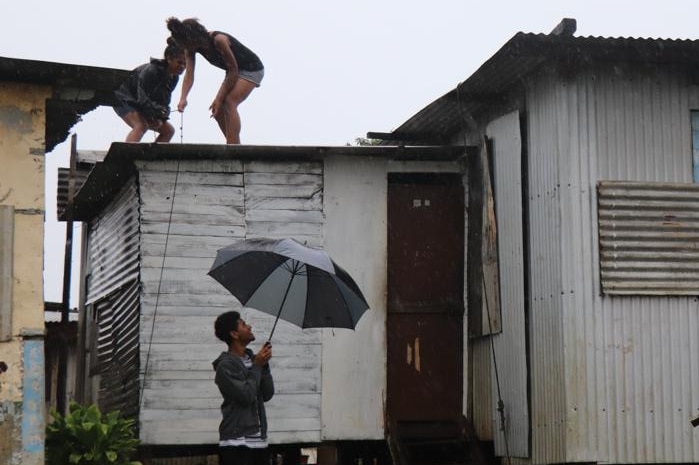 Two girls stand on the tin roof of a building in the rain, while a man below holds an umbrella.