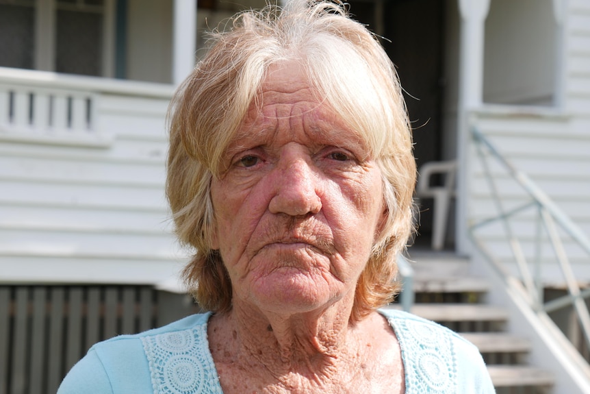 An older woman with fair hair standing out the front of her house.