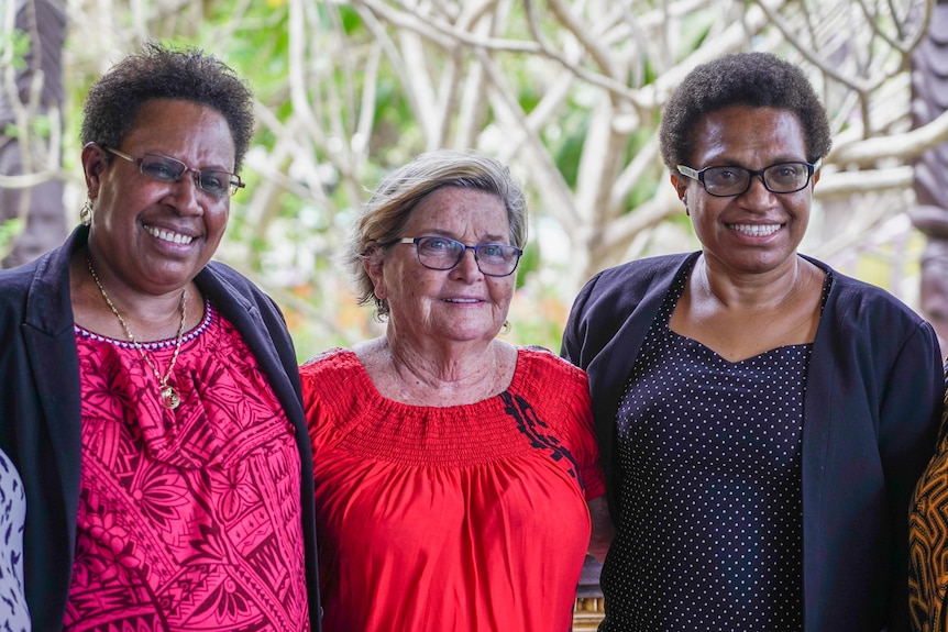 Three women stand together, in brightly coloured clothes, smiling off camera