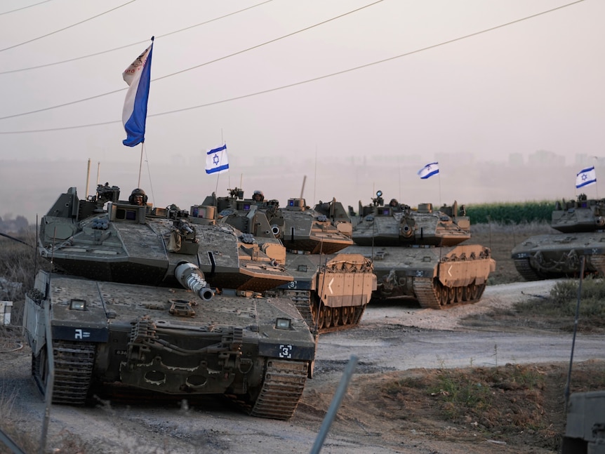 Four Israeli tanks drive in a line, with small Israeli flags flying from them.