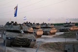 Four Israeli tanks drive in a line, with small Israeli flags flying from them.