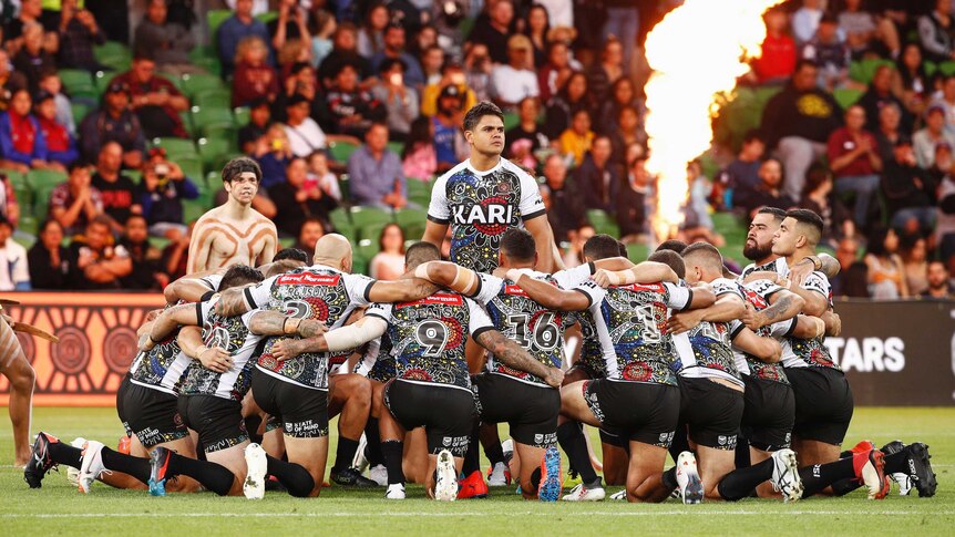 A group of NRL players kneel around Latrell Mitchell with a flame in the background