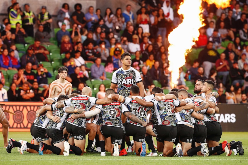 A group of NRL players kneel around Latrell Mitchell with a flame in the background