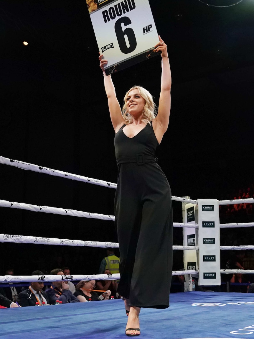 A blonde woman in a black cocktail dress holds up a 'round 6' sign in the ring during a boxing bout