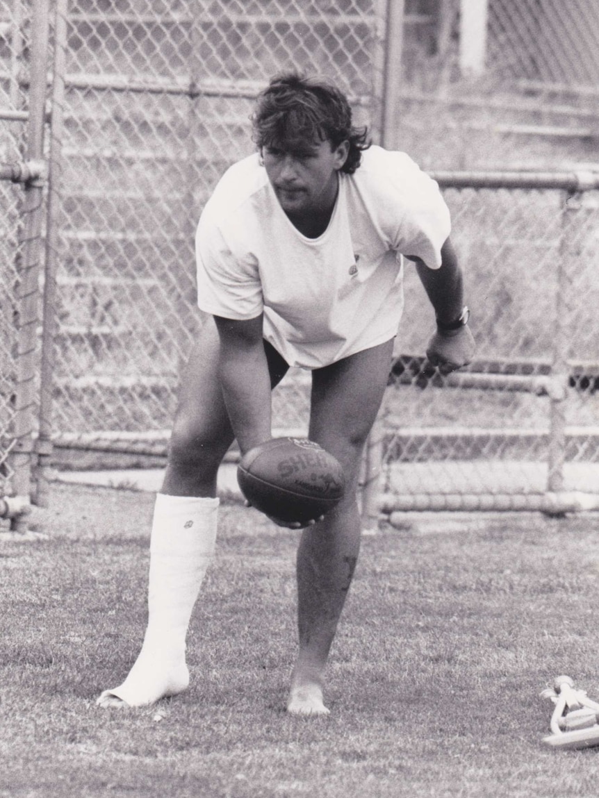 A man with a cast up to his knee prepares to handball a Sherrin football.