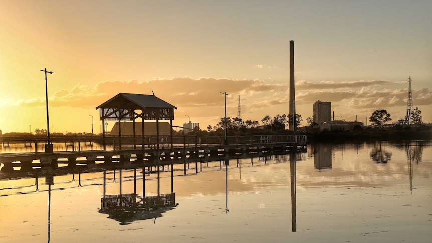 A golden sunset reflects a large industrial site onto still water at dusk.