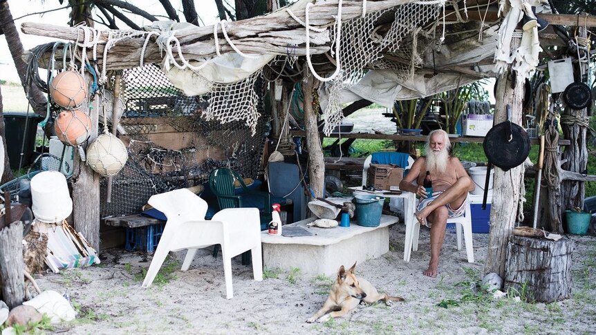 In a make-shift shelter of driftwood, netting and old buoys, a white-haired man sits in a plastic chair holding bottle of beer.