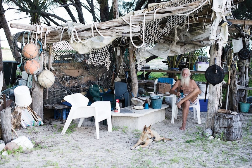 In a make-shift shelter of driftwood, netting and old buoys, a white-haired man sits in a plastic chair holding bottle of beer.