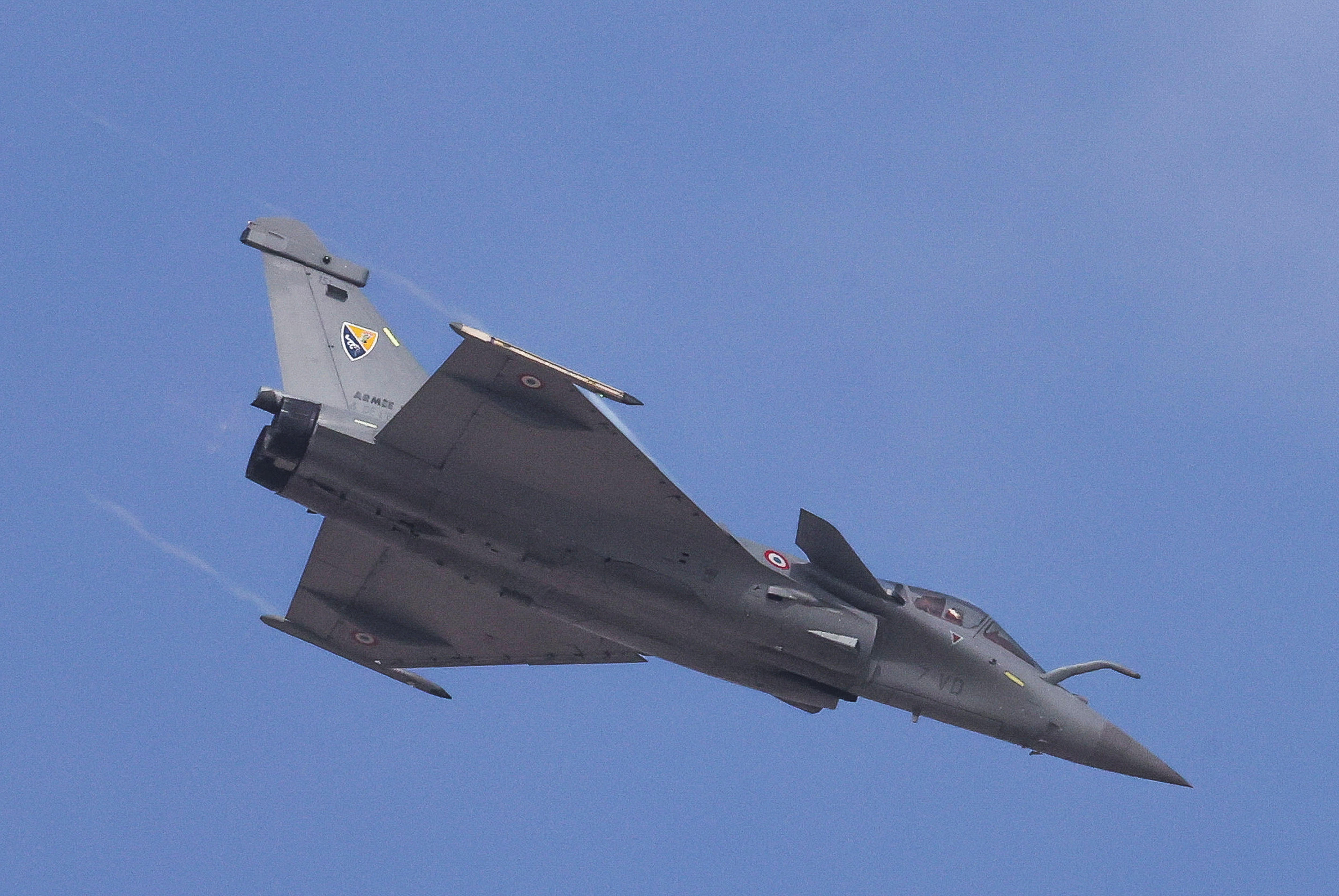 A grey fighter jet flying against a blue sky, seen from below.