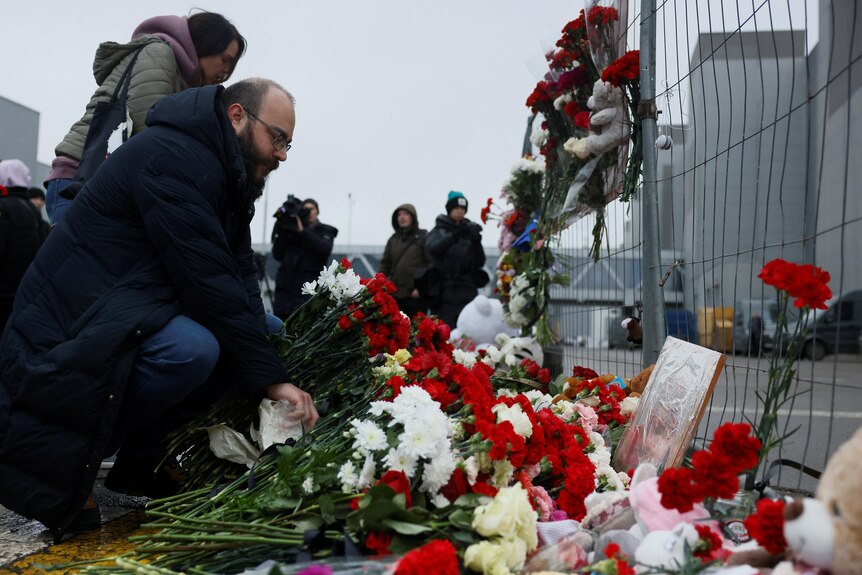 A man lays red flowers near a fence as other mourners watch on in the background