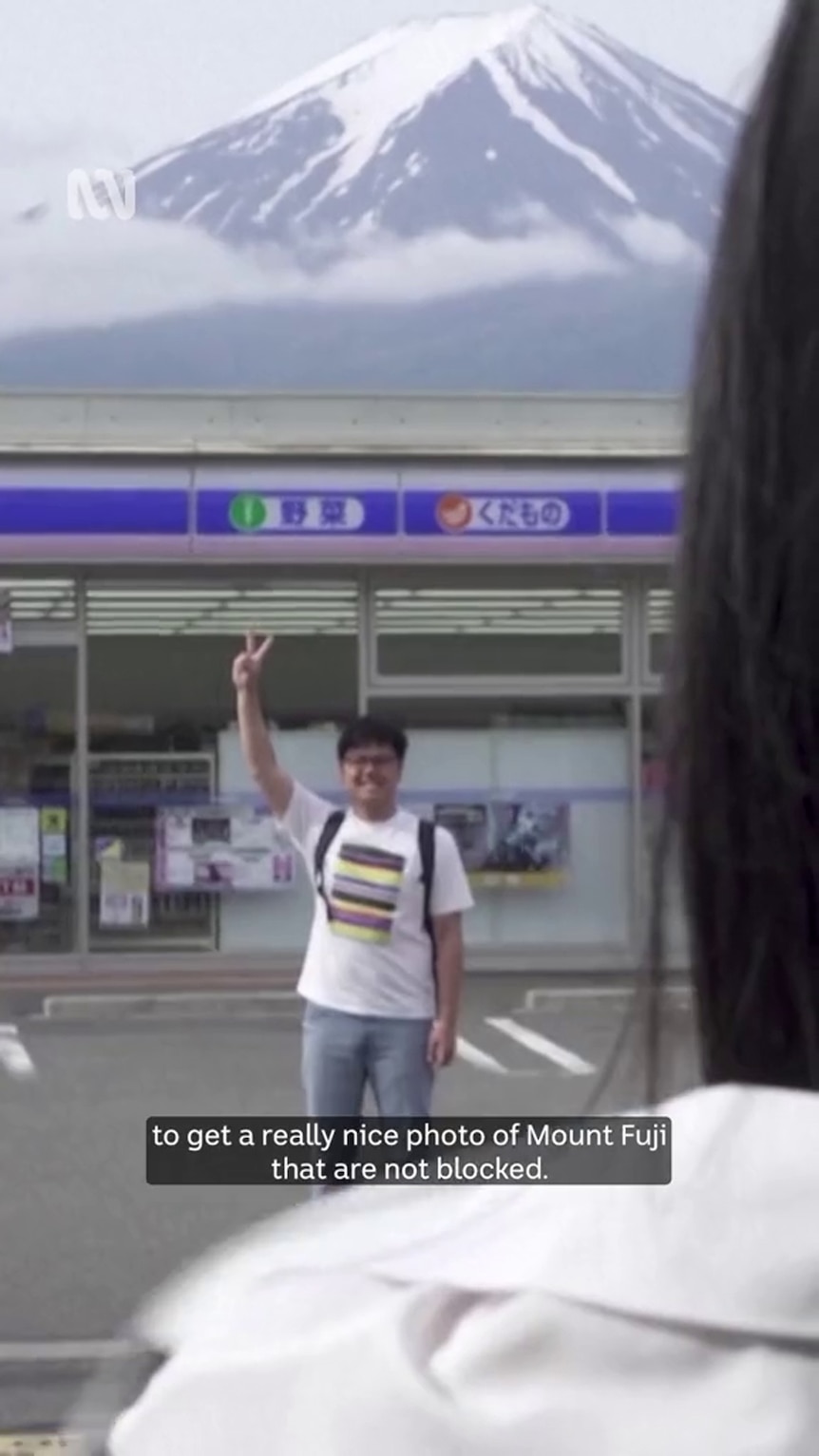 An Asian man flashes the peace sign in front of a storefront with a prominent snow-capped mountain visible in the background