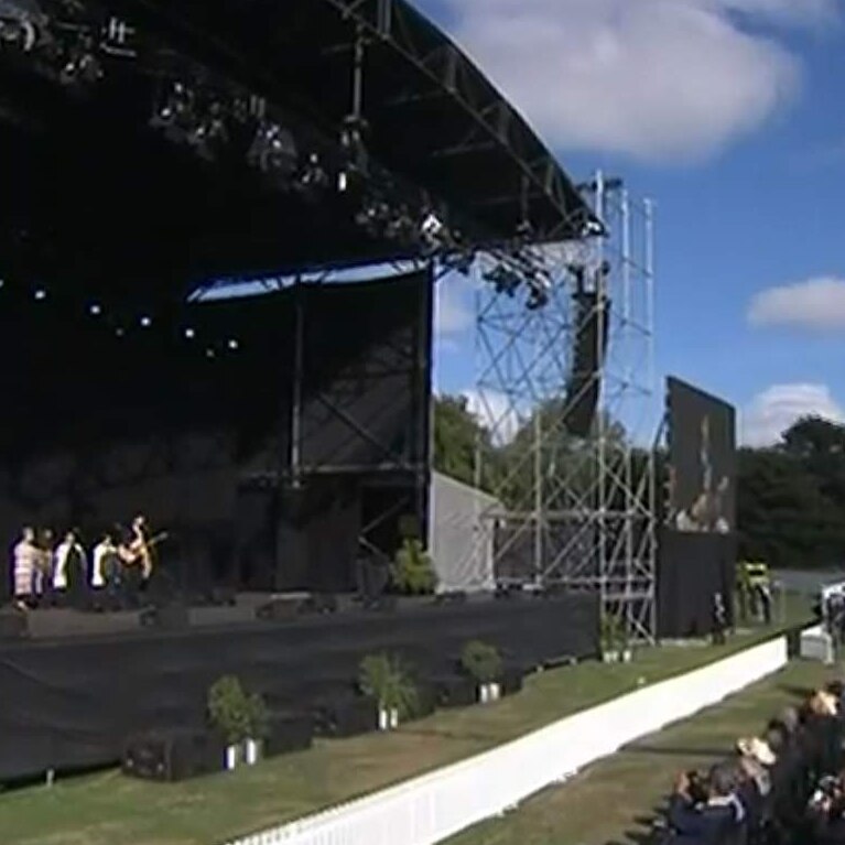 A stage features performers and officials. To the right is a large audience of mourners