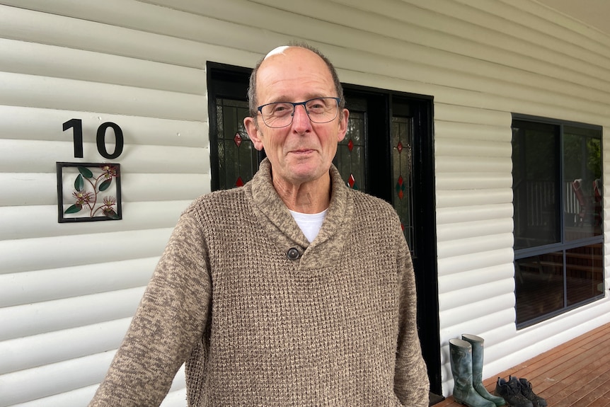 A balding man wearing a brown sweater and glasses, standing in front of a weatherboard house