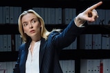 A young, smartly dressed blonde woman points one hand in the air. Legal folders line the wall behind her.