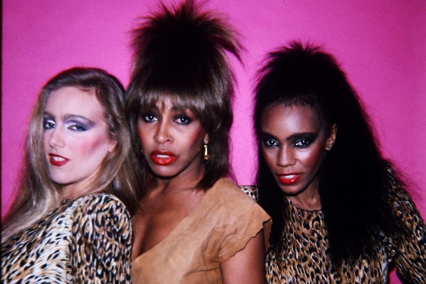 The singer Tina Turner with iconic 80s hair surrounded by two female dancers in leopard print, pink background