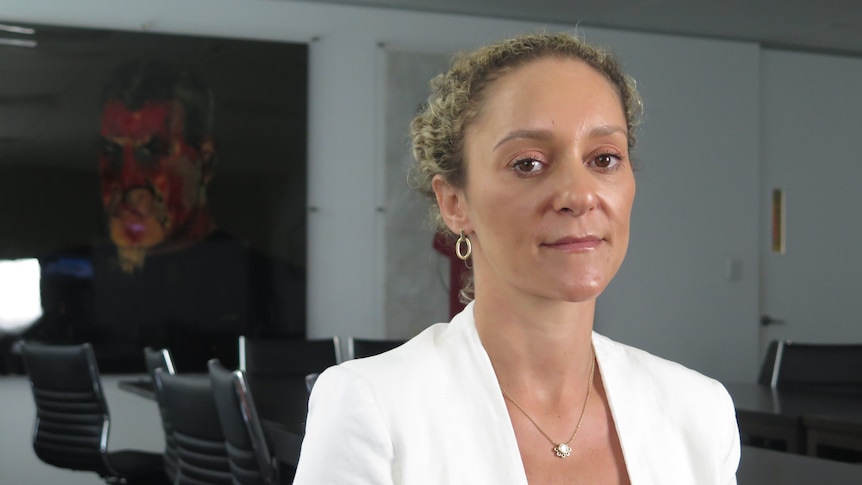 A women with blonde/brown hair and a white suit, standing in a boardroom