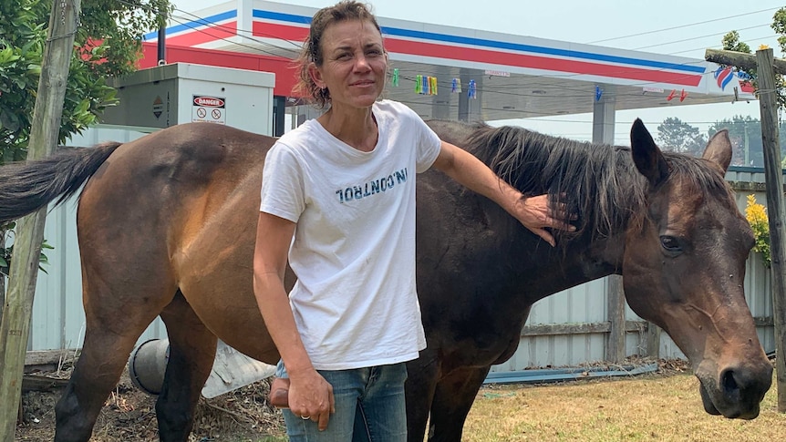 A woman in a white t-shirt stands near a petrol station with a horse.
