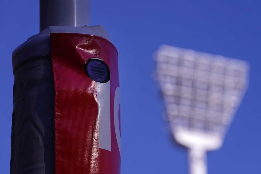 A small, round camera can be seen inside the padding around a goalpost. A floodlight is in the background