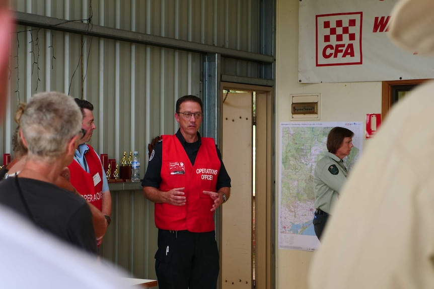 A man in a high-vis vest is speaking in front of a handful of people inside a fire brigade garage.