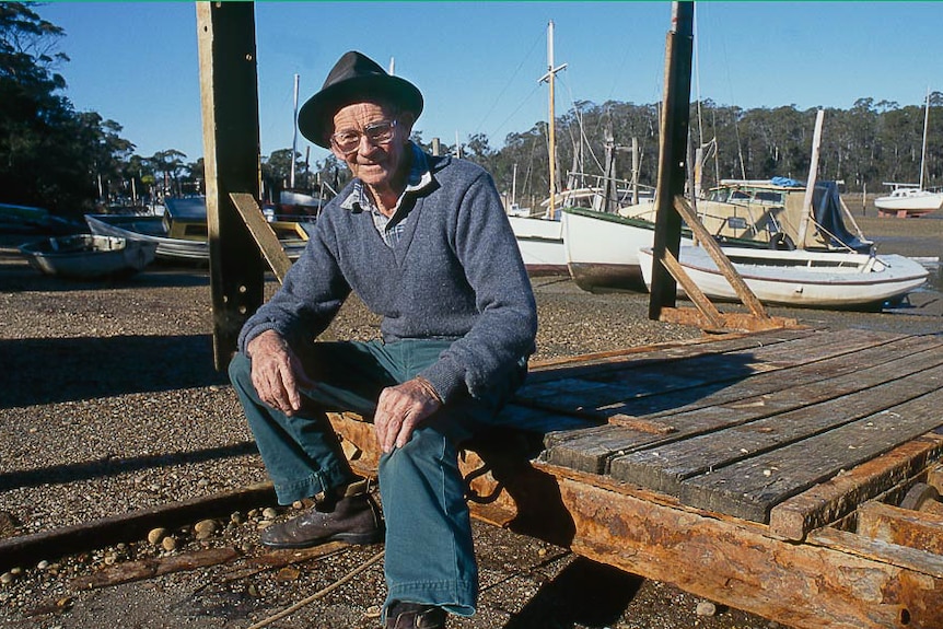 Old man in a hat sitting on a slipway trolley with many boats behind.
