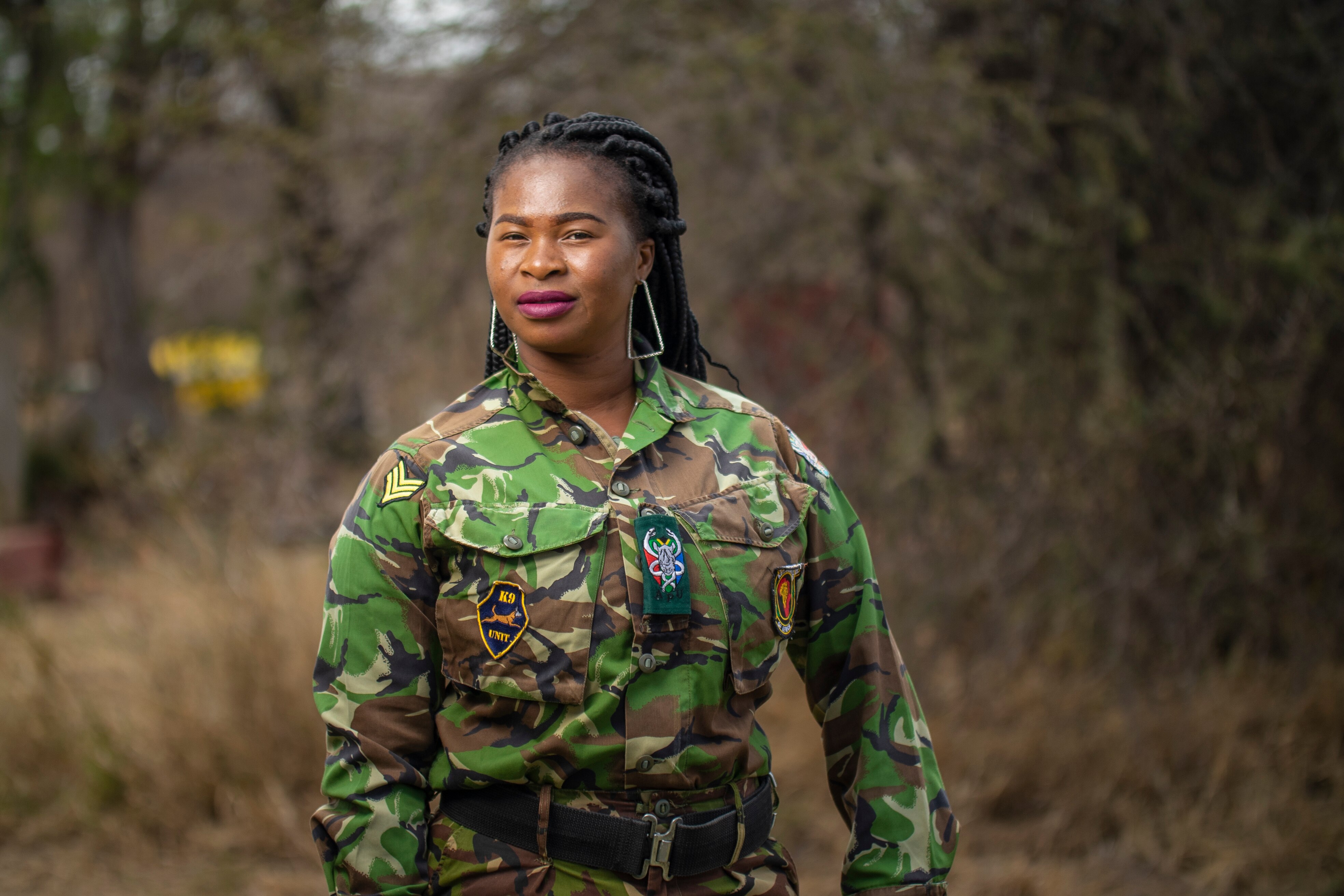 Portrait of a young African woman in a camouflage uniform in the bush. She's wearing purple lipstick and big earrings.