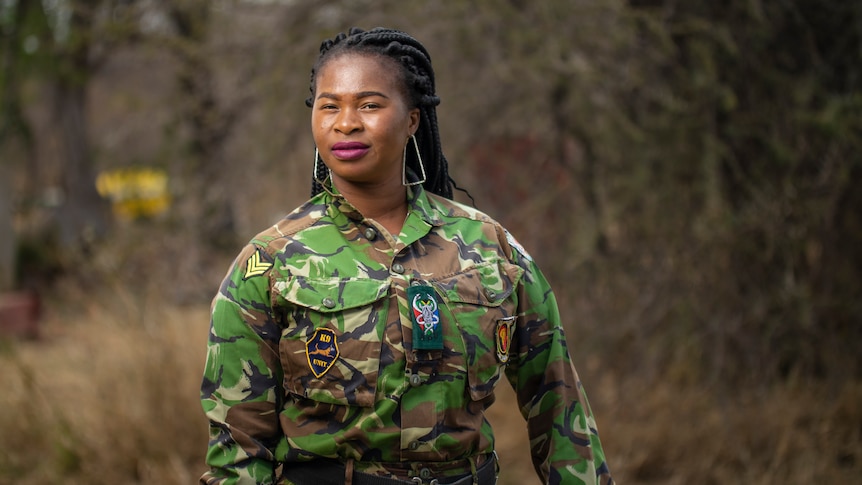 Portrait of a young African woman in a camouflage uniform in the bush. She's wearing purple lipstick and big earrings.