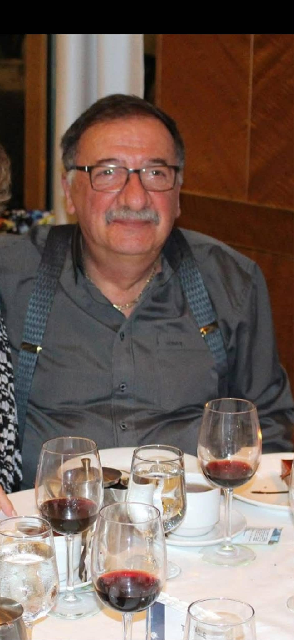 Un hombre vestido con una camisa gris oscuro, tirantes azules y gafas con montura oscura se sienta en una mesa con copas de vino y platos.