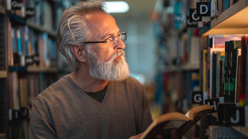 An older man looks at the shelves in a library while holding an open book