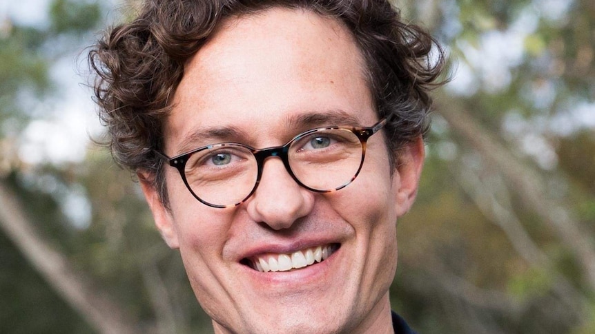A Caucasian man with blue eyes, dark curly hair and glasses, smiles at the camera in a professional headshot