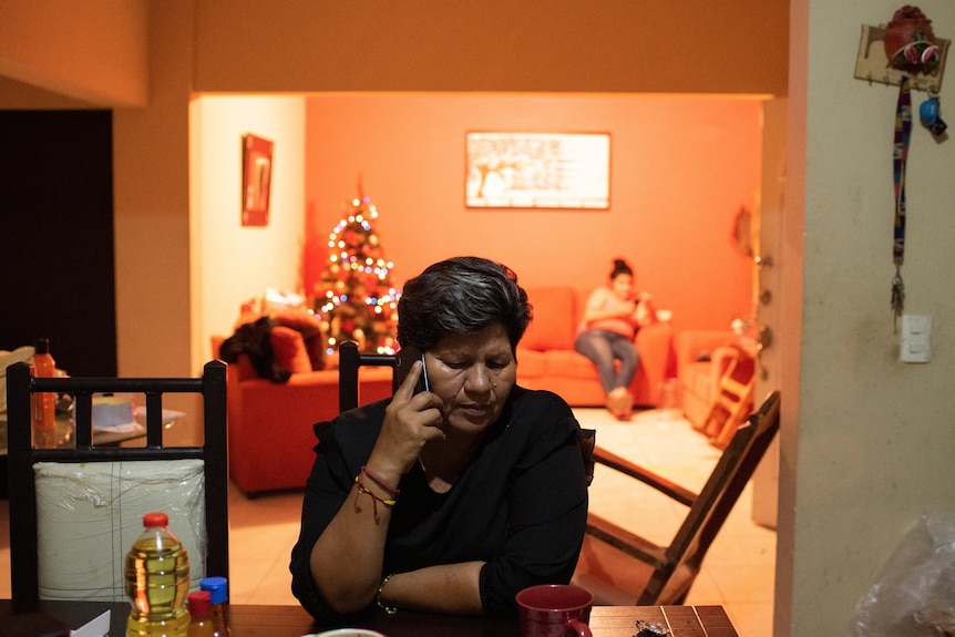 A woman is pictured sitting at a table while on the phone. Behind her, someone is sitting on a bright orange couch.
