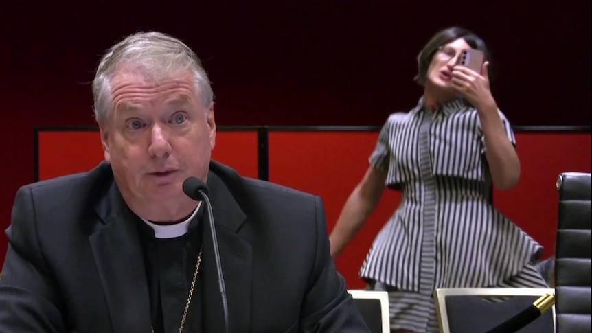 A bishop is speaking into a mic as a woman behind him holds a phone and interrupts him