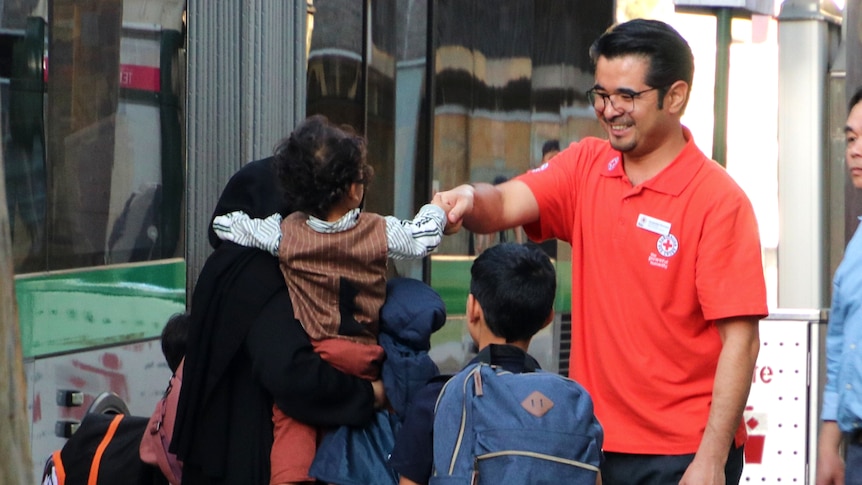 A man wearing a Red Cross shirt greets people as they step off a bus