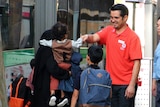 A man wearing a Red Cross shirt greets people as they step off a bus