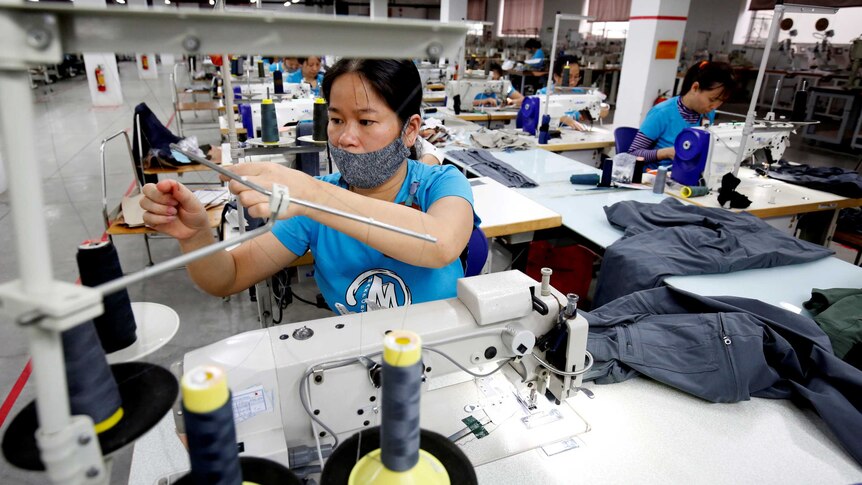 A woman works at a sewing machine. Other workers are visible behind her. She is wearing a mask on her face.