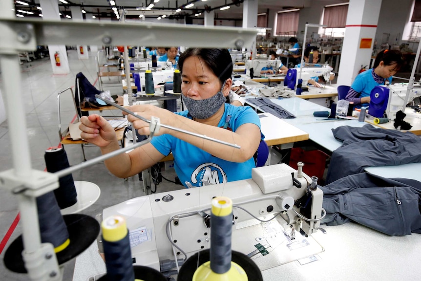 Una mujer trabaja en una máquina de coser.  Detrás de ella se ven otros trabajadores.  Lleva una máscara en la cara.