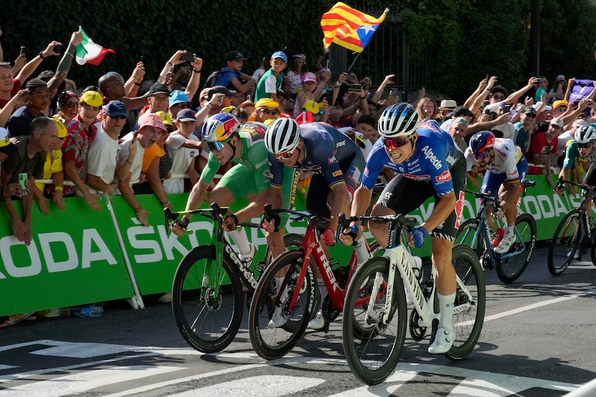 A cyclist grits his teeth as he races over the line in front in a sprint finish, with two other riders to the left of him. 