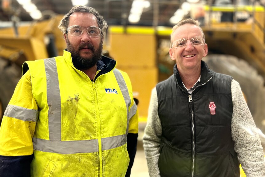 A man wearing a dirty bright yellow jacket stands next to another man wearing a blue vest inside a truck mechanic workshop.